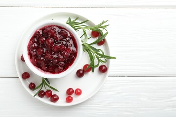 Tasty cranberry sauce in bowl, berries and rosemary on white wooden table, top view. Space for text