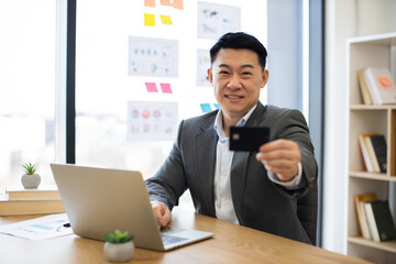 Smiling Asian middle-aged businessman sitting at desk using laptop, holding credit card for online payment in modern office. Concept of e-commerce, technology, digital transactions.