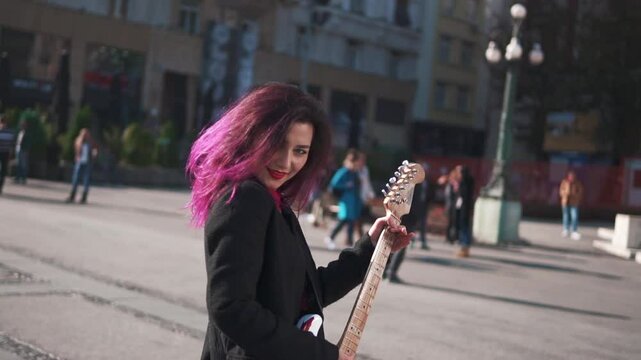 Music lifestyle concept. Daring rebel woman with purple hair playing electric guitar at city center during bright sunset. People walking in background. Asian girl rock star, provocative behavior.