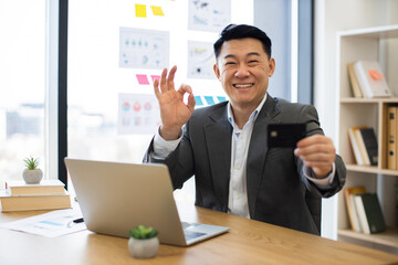 Asian businessman in office makes online payment with credit card. Smiling, he gives approval gesture while using laptop, suggesting success and satisfaction.