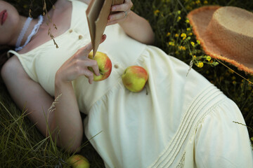 Woman,  lying in the green grass and reading a book, dressed in a beautiful yellow dress, holding an apple in the hand, enjoying rest.