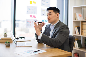 Asian businessman recording audio message with smartphone while sitting at modern office desk. Professional wearing formal suit engaged in digital communication utilizing technology for efficient work