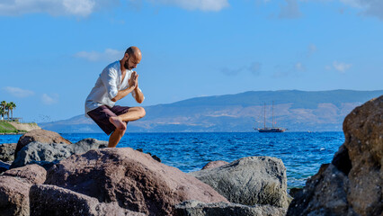 A middle-aged charismatic man, wearing a white shirt, is relaxing and enjoying the sun by doing yoga poses on the rocks by the seaside on a sunny summer day