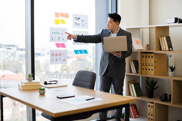 Asian businessman in professional suit analyzing charts and graphs during video call. Standing by panoramic window with laptop, pointing at data visuals. Modern office setting with urban view.