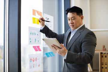 Asian male businessman in office works with graphs and charts on panoramic window using tablet. Professionally dressed in suit, he focuses on analyzing data, showcasing modern business environment.