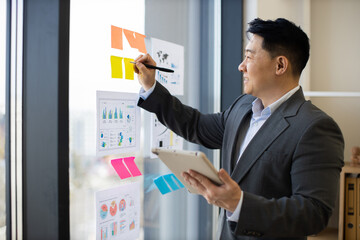 Asian businessman in formal suit analyzing graphs and charts on panoramic window using digital tablet. He is focused on data visualization and office productivity.