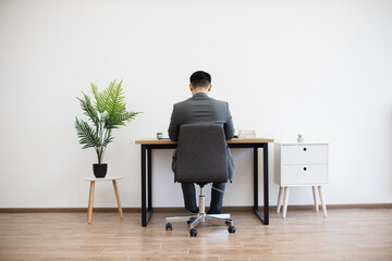 Asian male businessman sitting at desk working on laptop in modern minimalist office setting....