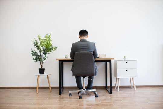 Asian male businessman sitting at desk working on laptop in modern minimalist office setting. Office includes white walls, potted plant, and wooden furniture, creating a clean workspace atmosphere.