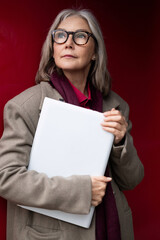 Stylish elderly woman with glasses confidently holding a folder against a striking red backdrop in an urban setting