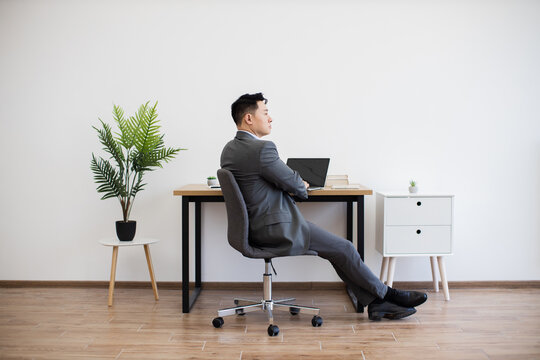 Asian man in business attire sitting in office chair, relaxing in modern workspace. Minimalist office features wooden desk, laptop, indoor plant on white wall backdrop. - Powered by Adobe