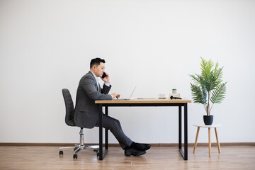 Asian businessman sitting at modern desk using laptop and making phone call. White wall background...