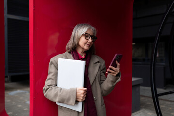 A senior businesswoman stands outside, engaged with her smartphone while holding a laptop under a...