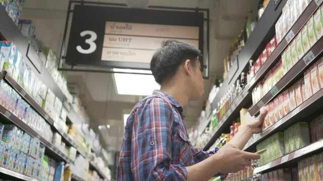 A man is shopping in a store and is looking at the third shelf. The store is well-stocked with a variety of products, including a large selection of beverages