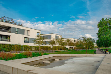 Modern apartment building surrounded by greenery under a clear blue sky. The residential complex features contemporary architecture with wooden accents, large windows, and balconies adorned with plant