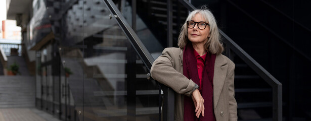 Middle-aged Caucasian businesswoman poses confidently at a modern urban office building on a sunny day