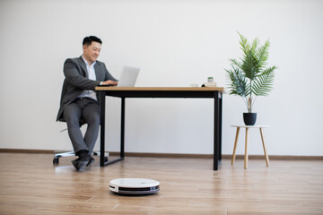 Asian businessman sitting at table using laptop in modern office space, while robot vacuum cleans floor. Scene depicts efficient, tech-savvy workspace with clean, minimalist design.
