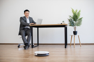 Asian businessman sits at desk working on laptop against minimalist white wall. Robot vacuum cleaner actively moves across wooden floor. Indoor plant adds greenery to modern office setting.