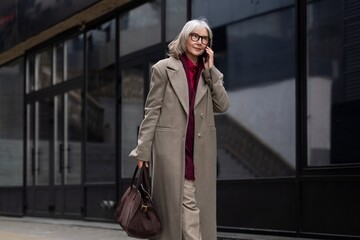Fototapeta premium Senior businesswoman dressed in formal attire walks outside while talking on her phone in an urban setting during daylight hours