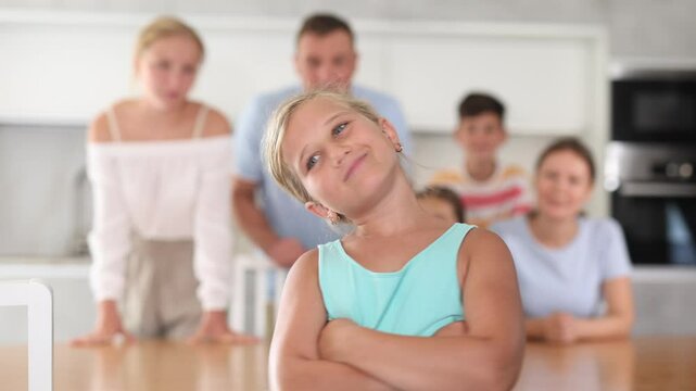 Offended pre-teen girl standing with arms crossed over chest at kitchen on background of her big family
