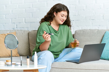 A young plus size woman with curly hair relaxes in her living room, sipping tea and trying...
