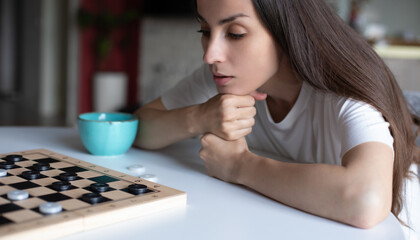 Woman in a white t-shirt intensely focuses on a checkers game, resting her chin on her hands. A teal mug sits beside her. The scene portrays deep concentration and strategy in a cozy indoor setting