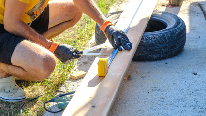A middle-aged man measuring with a tape measure to make repairs with lumber next to his boat in the garden of his house on a summer day