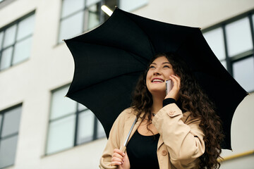 A joyful young plus size woman with curly hair chats on her phone under her umbrella during autumn...
