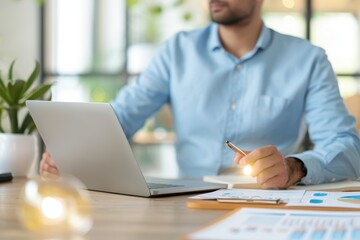Fototapeta premium Businessman working with laptop and financial documents in a bright office. Close-up of charts, graphs, and pen in a modern workspace.