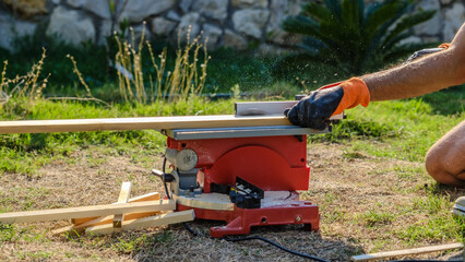 A middle-aged man cutting timber with a circular saw in the garden of his house on a summer day