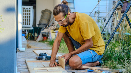A middle-aged man measures with a tape measure to make repairs with lumber in his garden on a summer day
