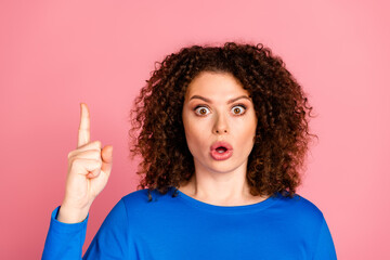 Surprised woman in blue sweatshirt with finger raised against pink background, conveying shock or...