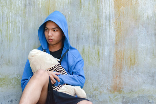 Asian teenboy in blue hoodie holding clothes doll and sitting on dirty floor and messy wall of an abandoned building, look sad, soft focus, concept for homeless, confinement and detention concept.