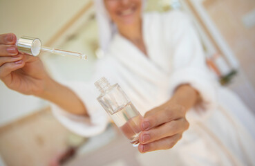 A woman in a bathrobe and towel on her head uses a dropper to apply skincare serum from a glass bottle. The bright bathroom setting suggests a fresh, self-care beauty routine and wellness lifestyle.