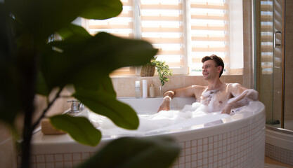 A man enjoys a relaxing bubble bath in a cozy, modern bathroom. Sunlight pours in through the window, while greenery and bath products surround him, creating a serene and refreshing atmosphere.