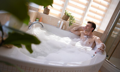 A man enjoys a bubble bath in a bright, modern bathroom. Surrounded by plants and bath products, he looks relaxed, leaning on the edge of the tub as sunlight streams in through the window blinds.