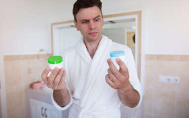 A man in a white bathrobe stands in a bathroom, holding two skincare containers and comparing them with a focused expression. The modern bathroom features tiled walls, conveying a self-care routine.