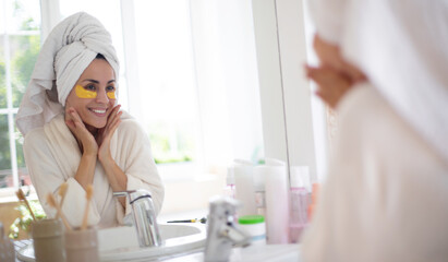 Woman in a bathrobe with a towel on her head smiles in the mirror while wearing golden under-eye patches. The scene highlights a beauty routine, skincare, and self-care in a bright bathroom setting.