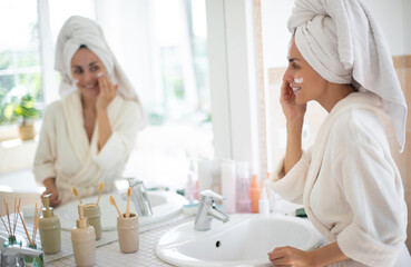 Smiling woman in white robe applies skincare cream while looking at herself in bathroom mirror. Her hair is wrapped in towel, surrounded by beauty products and plants, evoking fresh, relaxing vibe