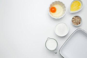 Empty white baking dish with ingredients for baking, including egg, milk, butter, sugar, and nuts on a white background. Flat lay composition for cooking preparation