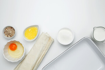 Near baking dish with ingredients, phyllo dough, egg, butter, nuts, and milk on a white background for baking Crinkle Cake