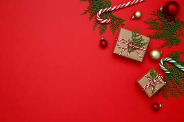 Gifts wrapped in craft paper, Christmas ornaments, thuja branches, and candy cane arranged on red background