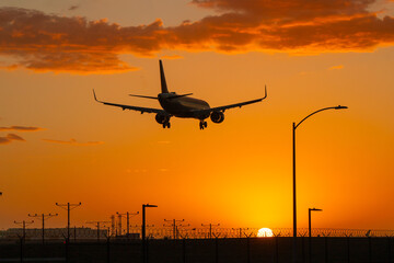 Airplane landing at Los Angeles International airport (LAX) at sunset. Orange sky and orange colored clouds. Airport infrastructure visible as dark silhouettes.