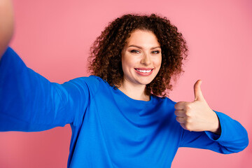 Cheerful young woman with curly hair giving thumbs up on pink background in casual blue sweatshirt