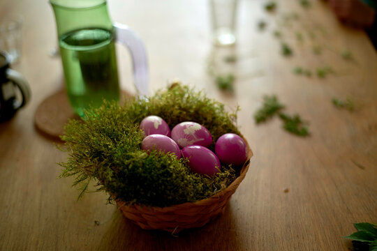 A wicker basket filled with pink painted eggs nestled in green moss on a messy wooden table
