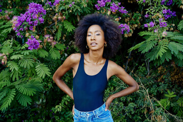 Confident woman with afro hair standing against a backdrop of vibrant purple flowers and lush green foliage.