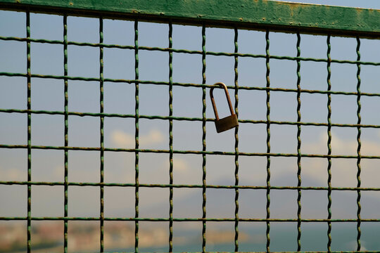 A rusty padlock hangs on a green metal grid fence with a blurred cityscape in the background, Naples, Italy