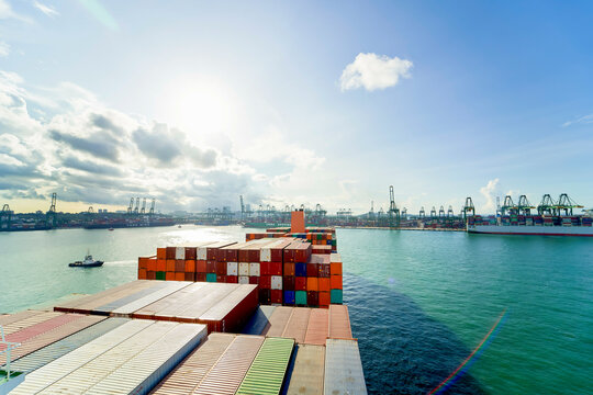 Cargo ship loaded with colorful containers heads out to sea on a clear day with a busy port in the background, Singapore