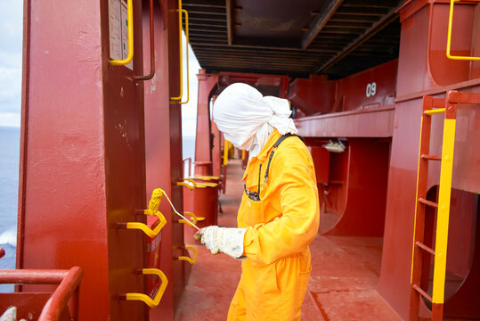 Red ship deck where a worker is painting the handles and ladders with yellow paint, his face covered in white fabric