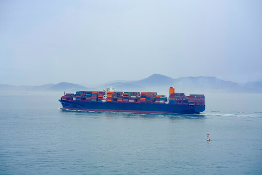 A cargo ship loaded with containers sails through misty waters with mountains faintly visible in the background, Busan, Korea