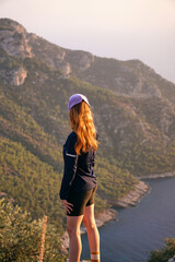 Naklejka premium A young red-haired girl stands in the mountains and looks at the sea. A sporty girl in the mountains